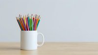 Assorted colored pencils arranged in white ceramic mug on wooden table against neutral background, suitable for education, creativity, art supplies, and design projects.