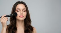 Young brunette woman applying makeup with brush, eyes closed, professional beauty routine for cosmetics and skincare advertising.