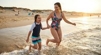 Happy mother and daughter running together through ocean waves on sandy beach during golden hour sunset creating joyful family vacation memories.