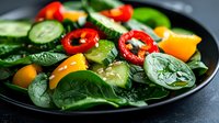 Fresh green salad with sliced cucumbers, red chili peppers, yellow bell peppers and baby spinach leaves on black plate, macro food photography.