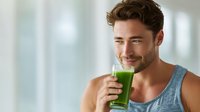 Young Caucasian man in gray tank top smiling while holding glass of green smoothie, representing healthy lifestyle and wellness routine.