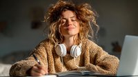 Young Caucasian woman with curly blonde hair wearing cozy sweater and headphones, writing in notebook while working from home office with laptop.