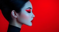 Asian woman with dramatic red eyeshadow and red lipstick in profile view against vibrant red background. Creative fashion makeup portrait showcasing bold artistic cosmetics.