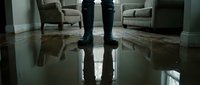 Person standing in rubber boots on flooded home interior floor, water damage disaster concept with furniture in background.
