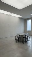 Modern minimalist interior with round metal table and black bar stools in empty concrete loft space, featuring geometric ceiling lights and large window for contemporary office or gallery design.