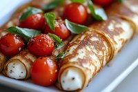 Rolled crepes filled with cream cheese served with roasted cherry tomatoes and fresh basil leaves on white plate, close up view of gourmet appetizer.