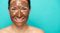 Young Hispanic woman with chocolate face mask smiling against turquoise background. Natural skincare routine, spa treatment, beauty wellness concept.