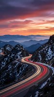 Winding mountain road with light trails at sunset showcasing dramatic orange and purple sky over snow-capped peaks and layered mountain ranges in distance.