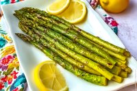 Grilled green asparagus spears garnished with lemon slices on white plate, close up view of roasted seasonal vegetables with bright citrus accents.