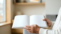 Senior adult with medium skin tone holding open blank white book with both hands, wearing white long-sleeve garment. Suitable for education, publishing, reading, and learning concepts.