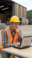 Senior construction worker in safety gear using laptop computer at industrial warehouse site for project management and planning operations.