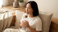 Young Hispanic woman relaxing on cozy beige sofa holding glass of water in bright modern living room with plants and natural home decor elements.