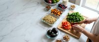 Hands preparing fresh salad with cherry tomatoes and greens on wooden cutting board surrounded by healthy ingredients including nuts, olives and legumes.