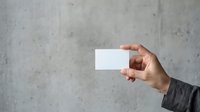 Hand of a man with medium skin tone wearing a dark sleeve holding a blank white business card against a concrete wall background for branding and corporate identity.