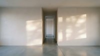 Empty modern hallway with white walls, natural light from windows casting geometric shadows on floor, leading to open doorway in minimalist interior space.