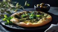 Freshly baked flatbread with aromatic herbs pesto, green mint leaves and olives on dark stone background. Mediterranean cuisine close up shot.