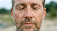 Middle-aged Caucasian man with closed eyes and beard in peaceful meditation pose outdoors with natural blurred background for wellness and mindfulness content.