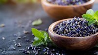 Dried lavender flowers in wooden bowls with fresh mint leaves on dark stone surface for aromatherapy, cooking, and natural wellness applications.