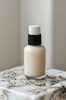 Glass bottle with beige liquid foundation or skincare serum on marble surface, minimalist cosmetic packaging with black cap, soft natural lighting.