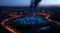 Aerial view of illuminated circular stadium with blue lights glowing at dusk, surrounded by city lights and roads creating leading lines through urban landscape at twilight.