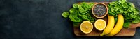Fresh ingredients for healthy green smoothie on wooden cutting board with spinach leaves, bananas, lemon halves and chia seeds in bowl on dark background.