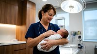 Smiling middle-aged Hispanic female nurse in scrubs gently holding a newborn baby in a modern hospital maternity ward.