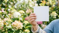 Young adult male with light skin tone holding blank white card in front of flowering garden background. Suitable for marketing, advertising, greeting cards, and promotional campaigns.