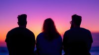 Silhouettes of three friends sitting together watching vibrant purple and orange sunset sky over ocean horizon, back view creating atmospheric mood scene.