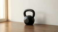 Black kettlebell weight on wooden floor against white wall, isolated fitness equipment for gym training, strength conditioning, and home workout routines.
