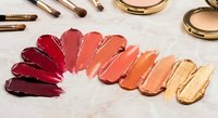 Colorful lipstick swatches arranged in gradient from deep burgundy to nude peach tones with golden makeup tools and compacts on marble surface.