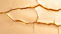 Cracked dry earth texture in warm beige and gold tones with dramatic lighting creating shadows along the fissures, abstract natural pattern background.
