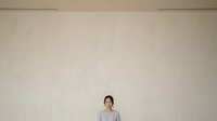Young Asian woman wearing casual gray shirt standing against plain beige wall, suitable for corporate, lifestyle, and portrait design projects.