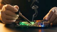 Professional technician repairing electronic circuit board with soldering iron and smoke rising from components in workshop setting.