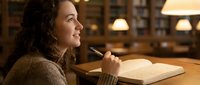 Young woman with curly hair studying in library, holding pen while reading book at wooden table with warm lighting for academic research and education.