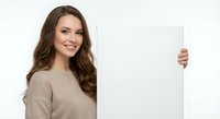 Young Caucasian woman with long brown wavy hair wearing beige sweater holding blank white poster board, smiling at camera on white background.