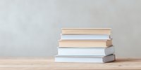 Stack of white and beige hardcover books arranged on wooden table against neutral wall background, creating minimalist composition for reading and education concepts.