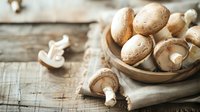 Fresh brown champignon mushrooms in rustic wooden bowl on vintage table with linen napkin, natural light and soft focus for food photography.