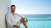 Middle-aged man with dark hair and beard, Mediterranean ethnicity, wearing white casual shirt, relaxing by infinity pool overlooking tropical ocean waters during daytime. Lifestyle and travel concept.