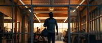 Professional businessman walking through modern office corridor during golden hour with colleagues working in background for corporate success concepts.