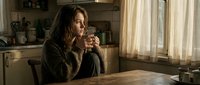 Young Caucasian woman in cozy knit sweater sitting at wooden kitchen table, holding coffee mug, gazing thoughtfully out window in warm morning light.