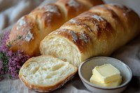 Fresh baked French bread loaf with golden crust, sliced to show soft interior crumb texture, served with butter pat in ceramic dish on linen cloth.