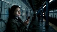 Man with dark hair and olive complexion wearing dark jacket holding smartphone in wet subway station at night, urban commuter checking mobile device in underground transit environment.