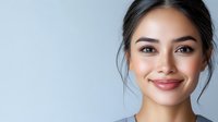 Young Asian woman with natural makeup and glowing skin smiling warmly against light blue background, hair pulled back in casual updo style for beauty and wellness concepts.