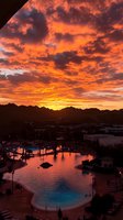 Dramatic orange and purple sunset sky reflects in resort swimming pool, with mountain silhouettes and palm trees creating serene evening atmosphere.
