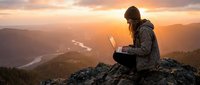 Woman working on laptop while sitting on mountain peak at sunset, overlooking valley with river and forest landscape below for remote work and digital nomad concepts.