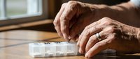 Elderly hands organizing daily medication in weekly pill dispenser on wooden table, healthcare management for senior adults at home.