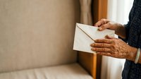 Senior woman with medium brown skin tone holding white envelope indoors, wearing dark sweater. Suitable for correspondence, mail, communication, and senior lifestyle concepts.