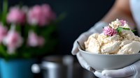 Creamy vanilla ice cream scoops in white bowl garnished with dried rose petals and mint leaves, elegant dessert styling against dark background with pink flowers.