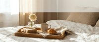 Cozy morning breakfast in bed with coffee cup and croissants on wooden tray in sunlit bedroom with white linens for hospitality and lifestyle concepts.