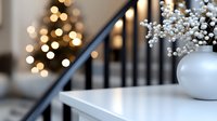 White baby's breath flowers in modern ceramic vase on white shelf against blurred Christmas tree lights and stair railing creating cozy winter home atmosphere.